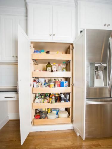 White kitchen pantry cabinet open, revealing pull-out shelves stocked with various food items, next to a stainless steel refrigerator.