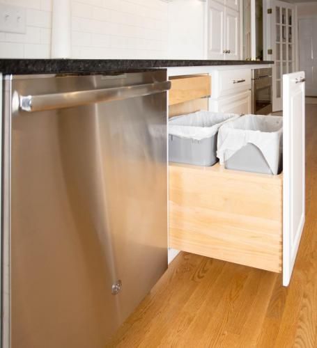 Stainless steel dishwasher next to a pull-out cabinet with two trash bins. White cabinet with wooden interior, on a hardwood floor.