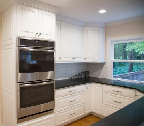 White kitchen with stainless steel double oven, white cabinets, and a dark countertop. A window overlooks a green outdoor area.