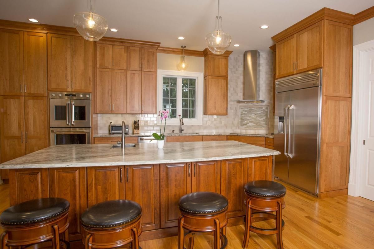 A well-lit kitchen with wooden cabinets, an island with stools, and stainless steel appliances. Light wood floors and a neutral backsplash complete the space.