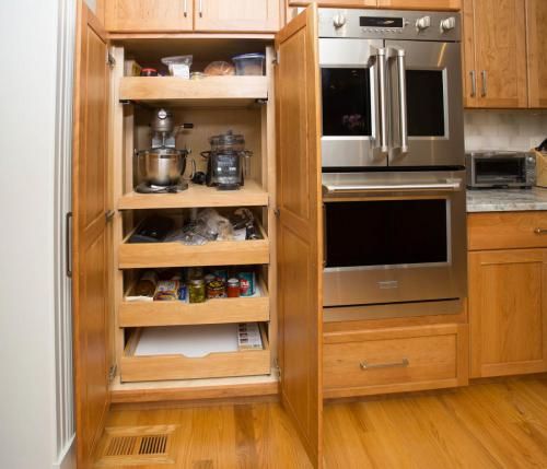 Kitchen cabinet with pull-out shelves displaying food and appliances, open beside a stainless steel double oven.