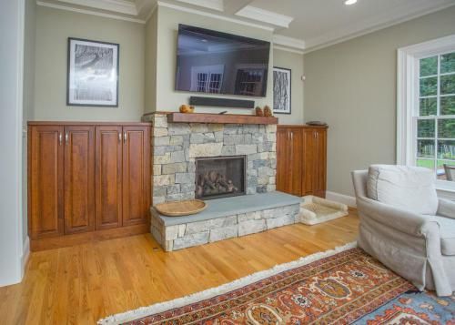 Living room with stone fireplace, built-in wood cabinets, and a mounted TV. Includes hardwood floors, a rug, and a white armchair.