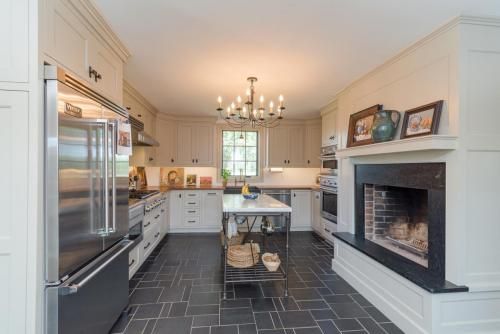 A kitchen with dark tile floors, light-colored cabinets, a stainless steel refrigerator, and a fireplace. An island with a marble top is in the center.