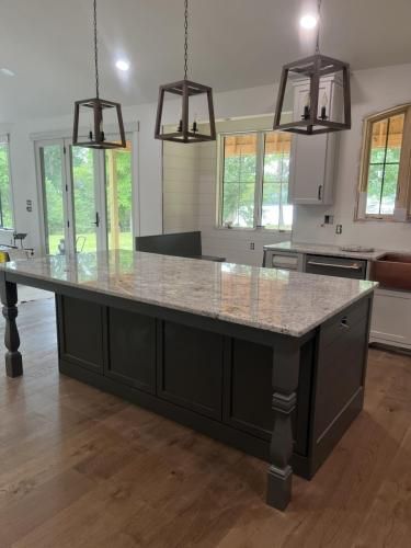 Kitchen island with granite countertop, dark gray cabinets, and three pendant lights above. Hardwood floors and large windows in the background.