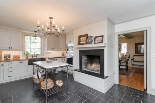 A kitchen with grey cabinets, black tile floor, and a marble-topped island. A fireplace is on the right, and a dining room is visible through the doorway.