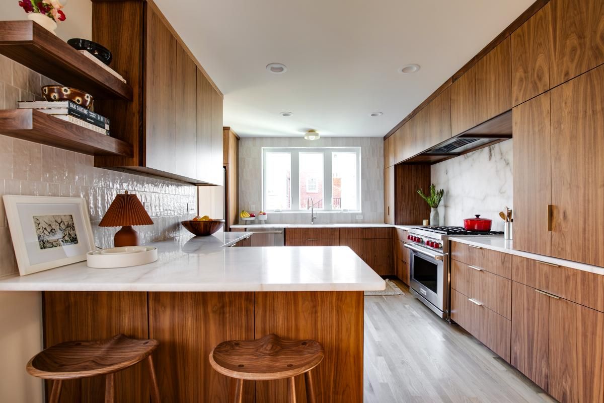 Modern kitchen with wood cabinets, white countertops, and a stainless steel oven. Two wooden stools sit at the counter.