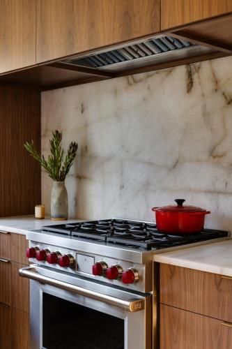 A kitchen with a stainless steel range, red knobs, and a red pot. The backsplash is white marble, and the cabinets are wood.