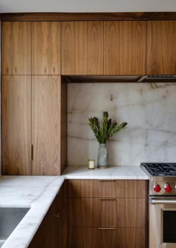A modern kitchen featuring wood cabinets, a marble countertop, and a stainless steel stove. A vase of green stems and a candle sit on the counter.