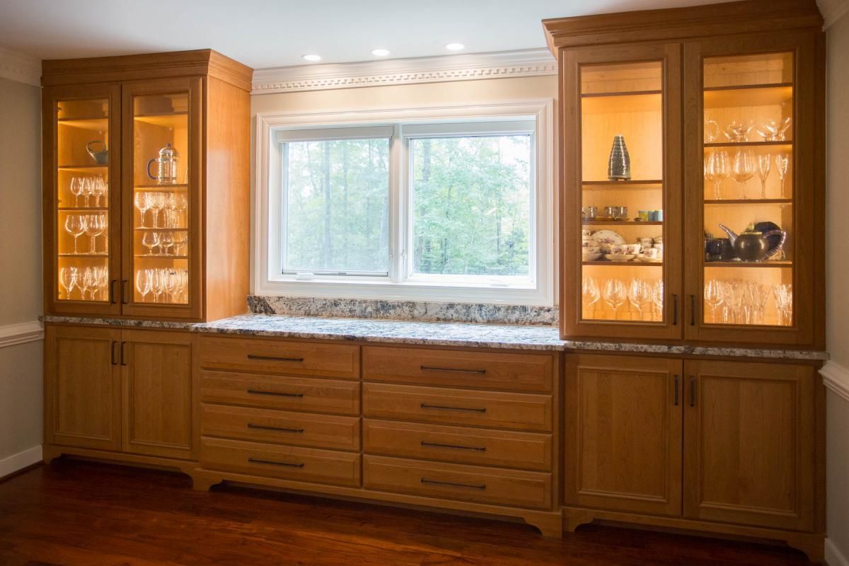 Built-in wooden cabinets with glass-fronted display cases flanking a window. Warm lighting illuminates glassware and decorative items.