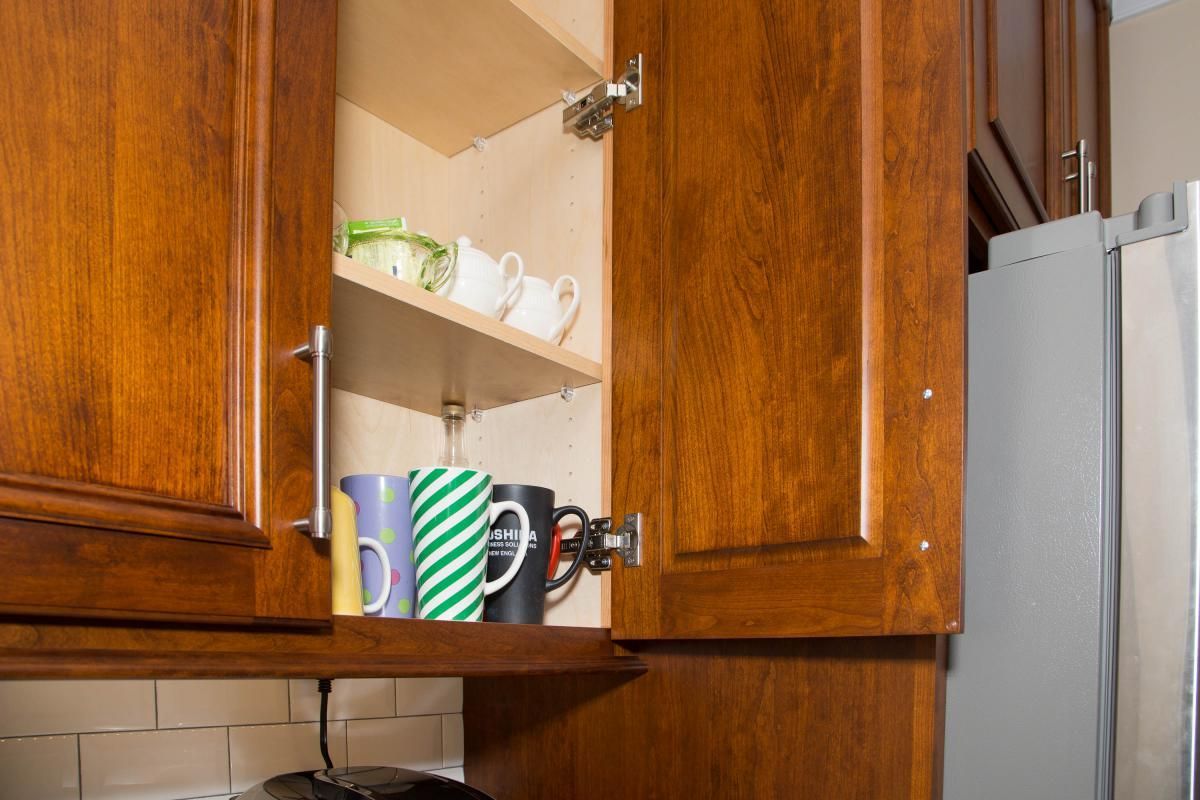Wooden kitchen cabinet open, filled with mugs on a shelf. The mugs are various colors and patterns.