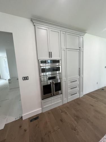 Built-in kitchen cabinets with stainless steel oven and refrigerator. Light gray cabinets with matching molding sit against a white wall and above hardwood floors.