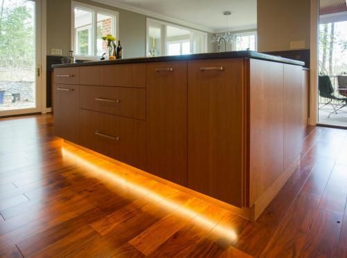 Kitchen island with brown cabinets and a black countertop, featuring under-cabinet lighting illuminating a hardwood floor.