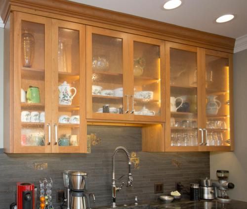 Light wood kitchen cabinets with glass doors, filled with dishware.  Backlit with warm lighting, above a sink and counter.