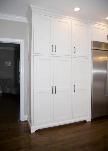 Tall white kitchen pantry cabinet with dark hardware, adjacent to a stainless steel refrigerator, set against a wood floor.