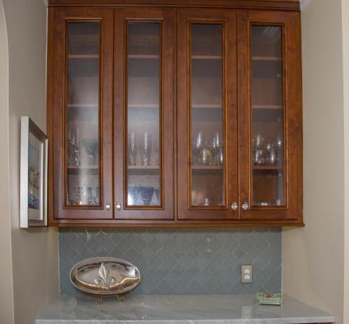 Wooden cabinets with glass doors above a counter with blue-gray tile backsplash, holding glassware. A silver tray rests on the counter.
