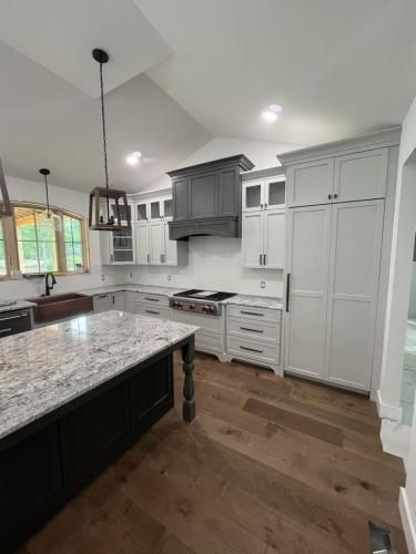 A modern kitchen with gray and white cabinets, granite countertops, and wooden flooring. A dark island sits in the center.