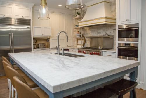 A spacious white kitchen with a marble countertop island, stainless steel appliances, and bar stools. Overhead lighting and a gold-accented range hood complete the elegant design.