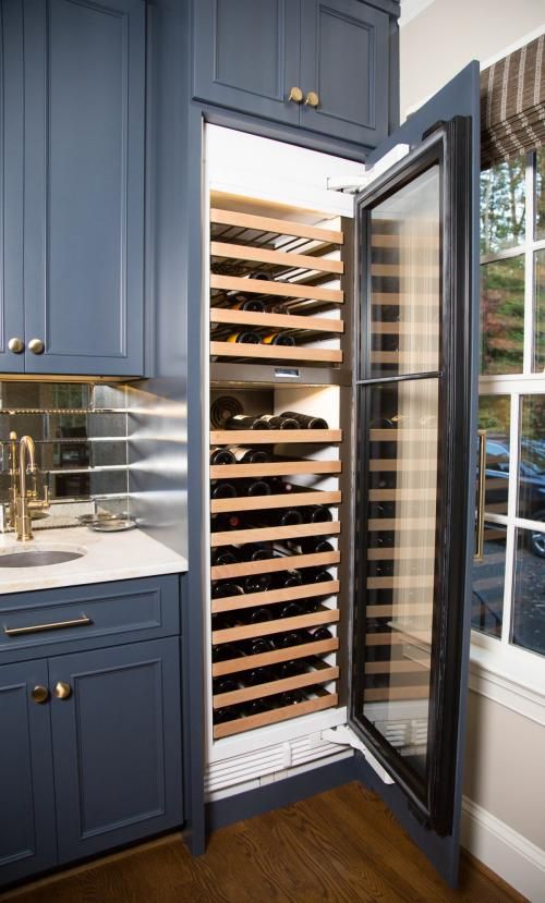 A built-in wine cooler in a blue cabinet, showcasing numerous bottles stored on wooden racks, with a glass door ajar.