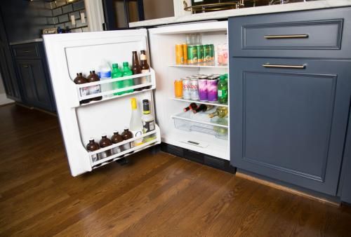 Mini-fridge built into kitchen cabinets, open and stocked with beverages and bottles on a hardwood floor.