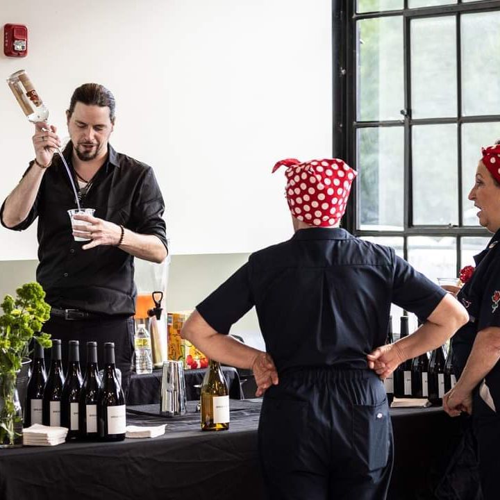 Bartender pouring a drink at a bar; two people watch. Dark shirts, red bandanas; bottles, drinks, and white wall visible.