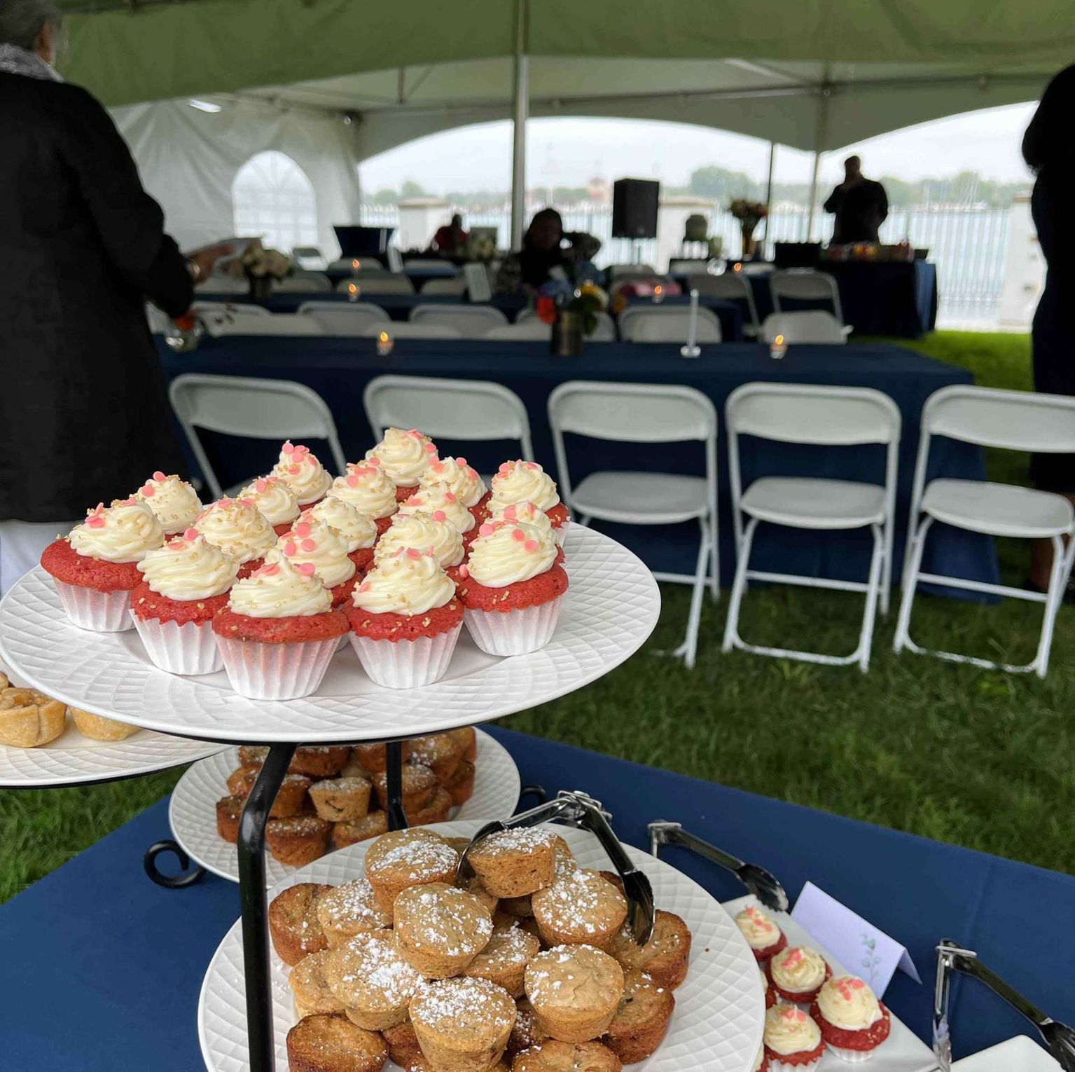 Cupcakes and mini muffins on tiered platters at an outdoor event with blue tables and white chairs under a tent.