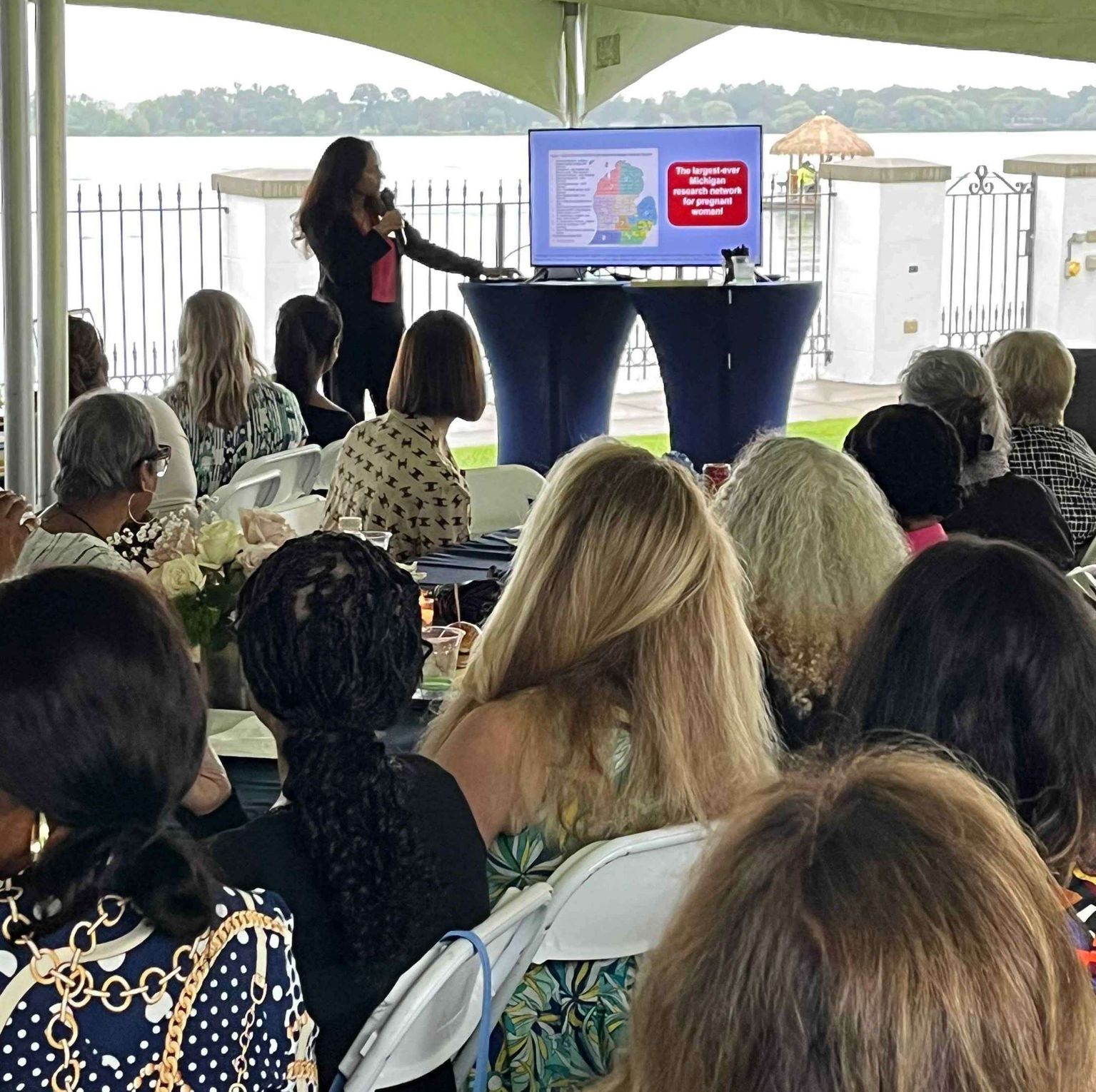 Woman presenting to an audience outdoors. She gestures towards a presentation on a screen. People sit at tables.