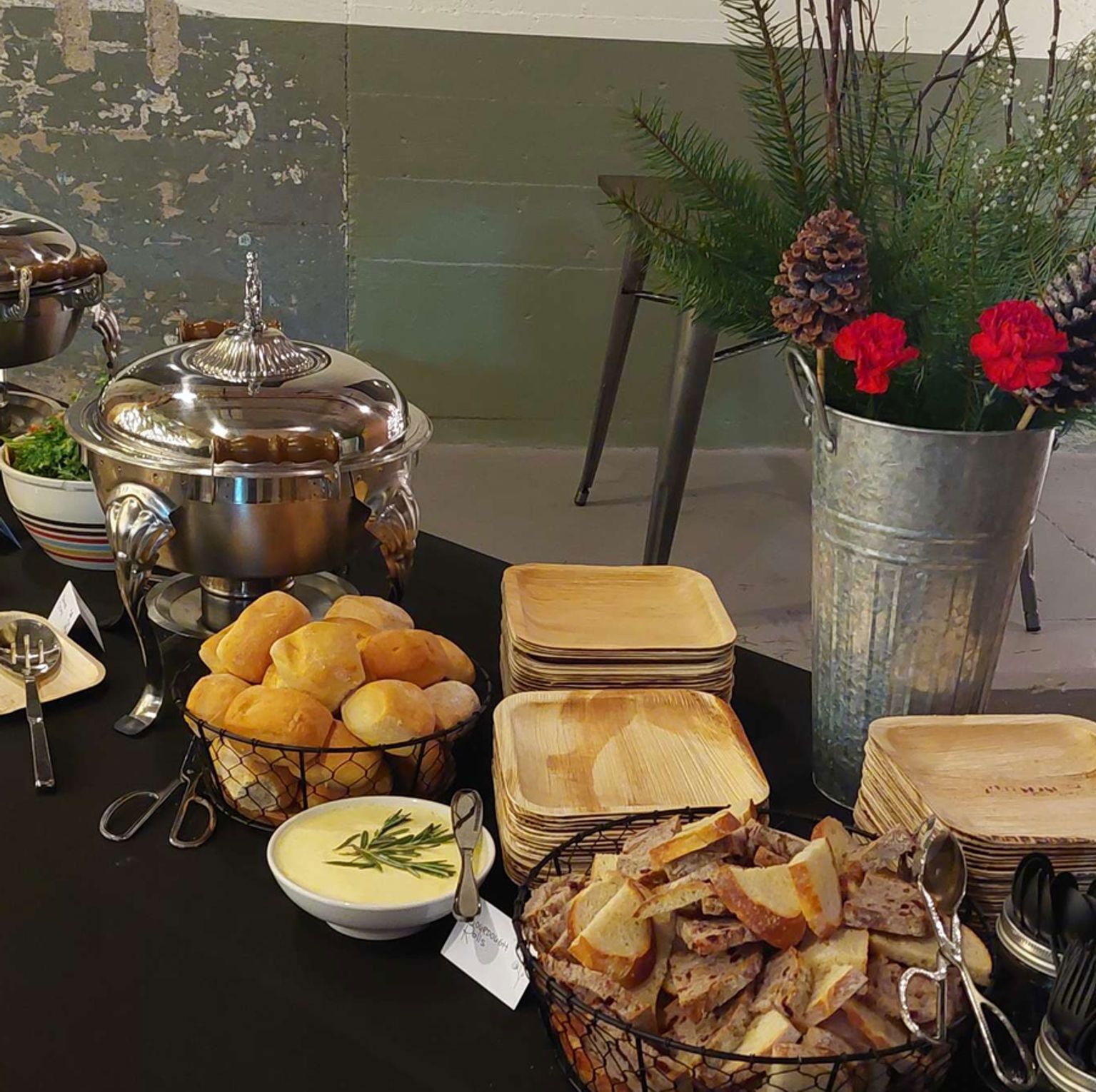 Buffet table with soup, bread rolls, bowls, plates, and a decorative vase with pine and flowers.