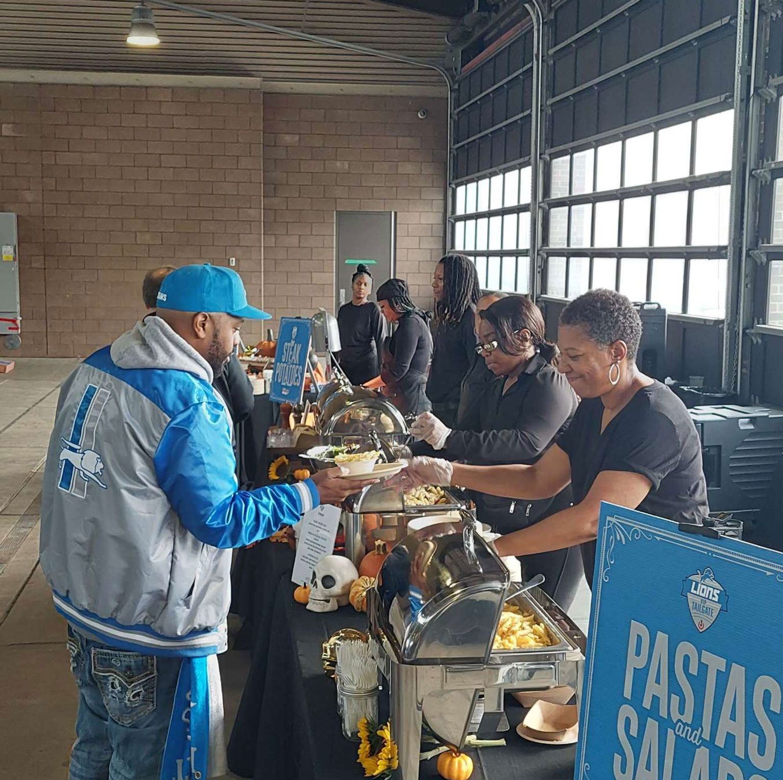 People serving food at a buffet. Man in Lions jacket getting a plate. Pasta salad sign, pumpkins, and a garage door.