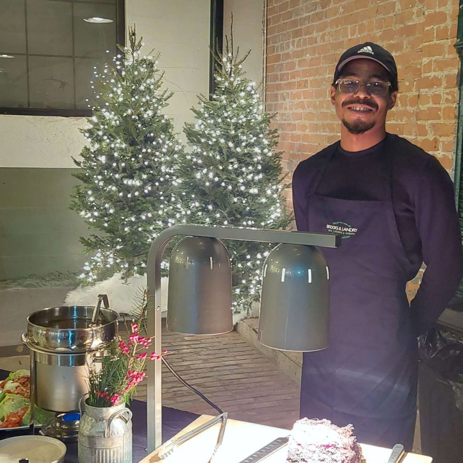 Man in apron smiles by food table; Christmas trees with lights in background.