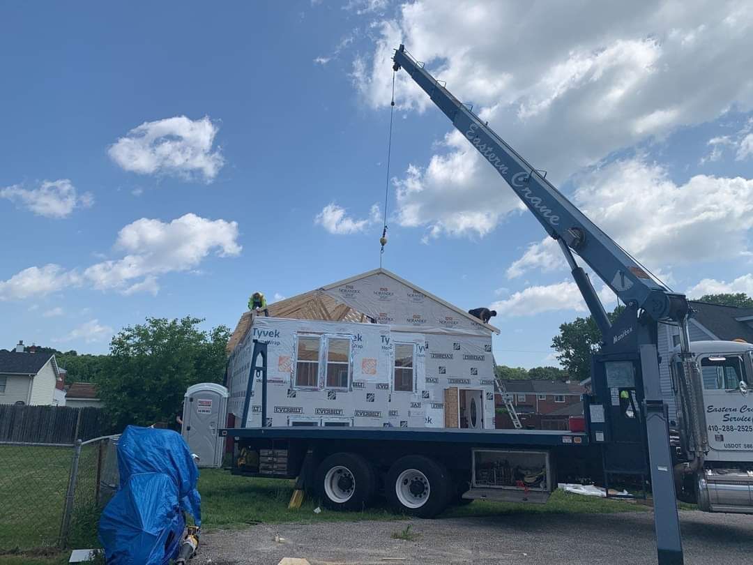 A crane lifts a small house onto a truck bed against a partly cloudy sky. Construction in progress.