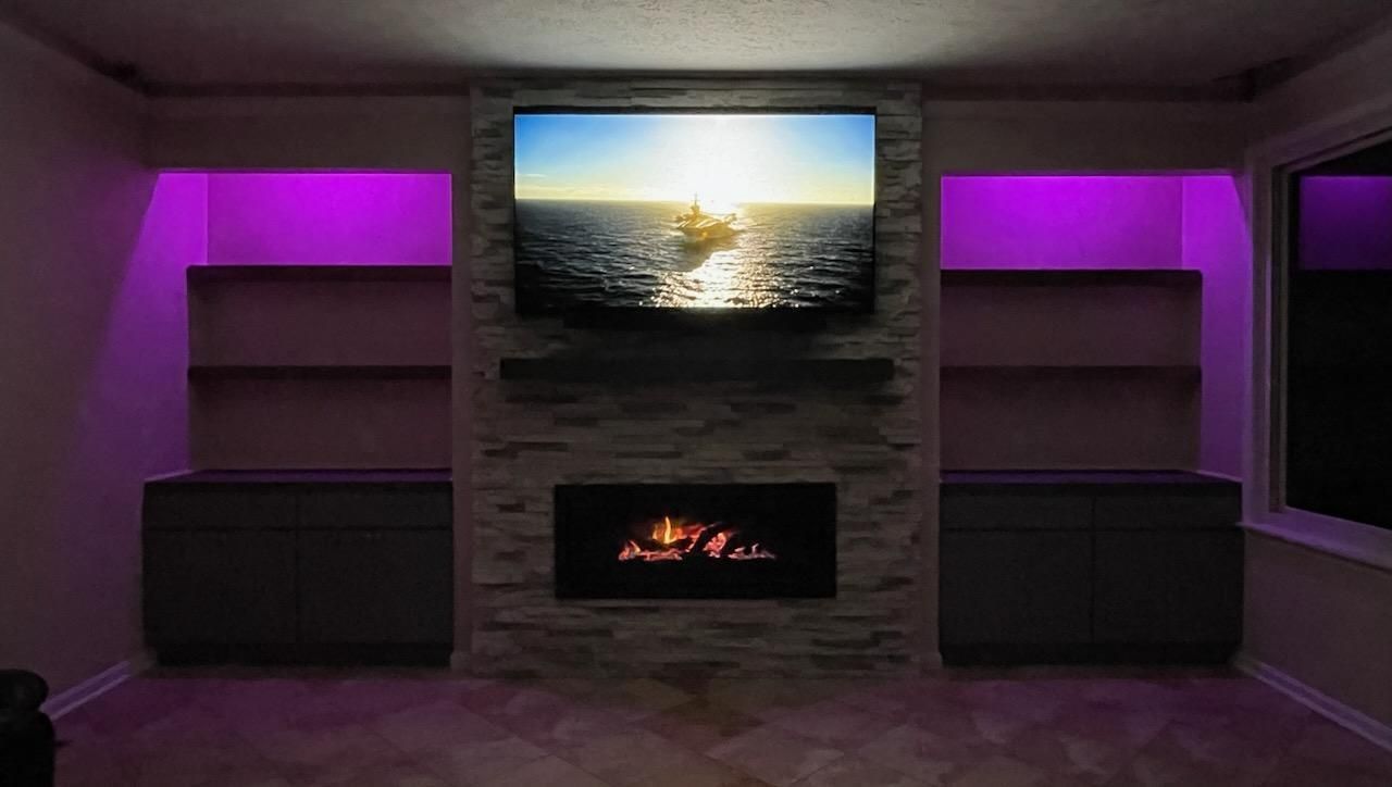 Room with fireplace, TV, and shelves, lit with purple accent lighting.