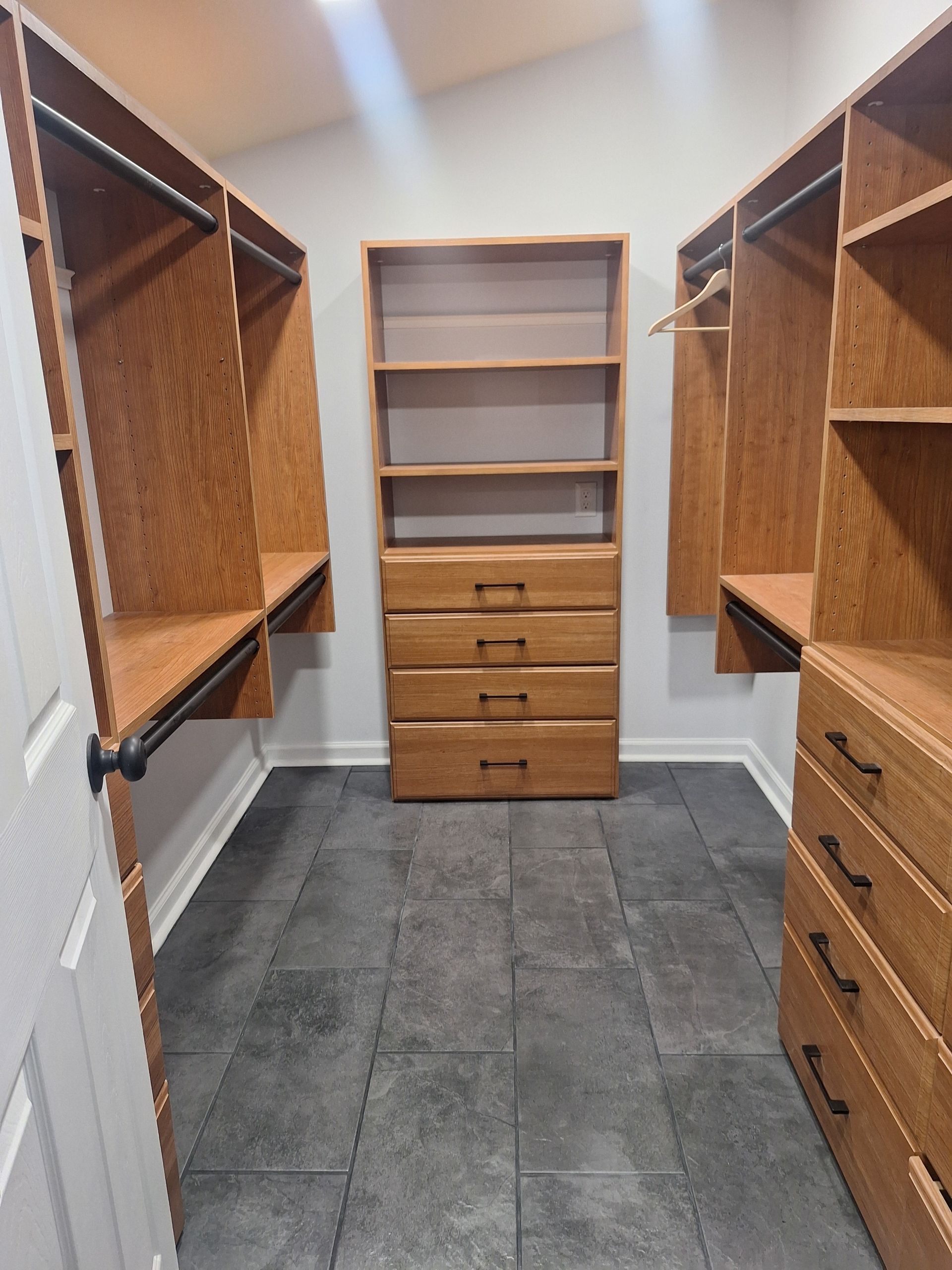 Walk-in closet with light wood shelving, drawers, and hanging rods. Gray tiled floor.