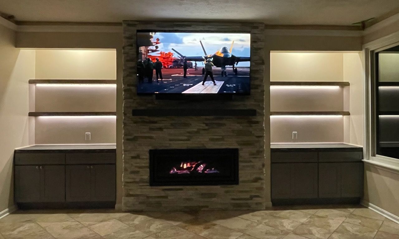 Built-in entertainment center with a fireplace, TV, and shelves. Beige walls, stone surround, and warm lighting.