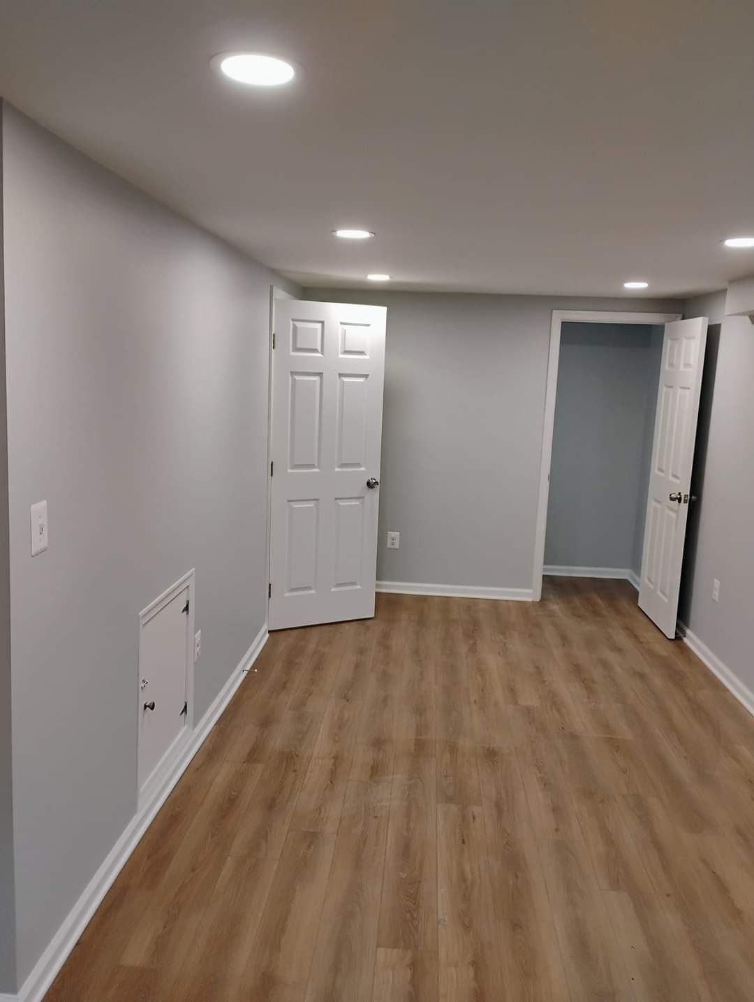 Empty room with gray walls, white trim, recessed lighting, and wood-look flooring; doors open.