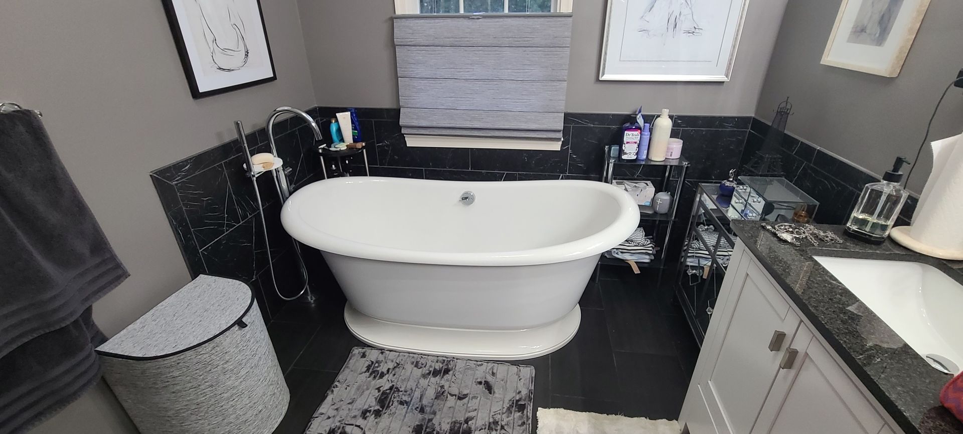 A bathroom with a white oval bathtub, black tile, gray walls, and a window with blinds.