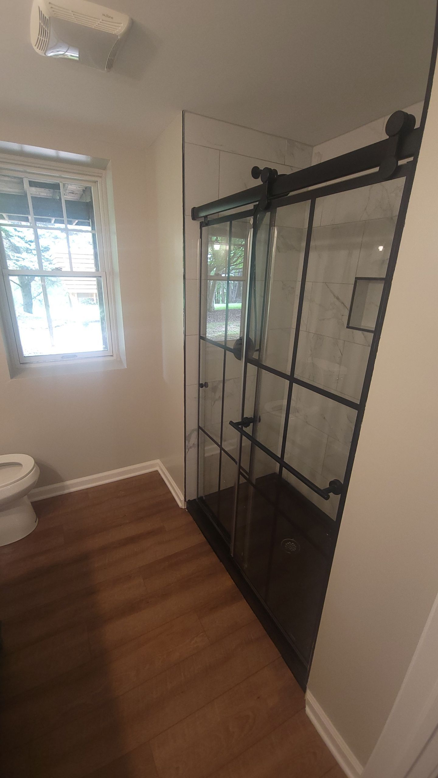Bathroom with a black-framed glass shower, toilet, and a window. Brown wood-look flooring.