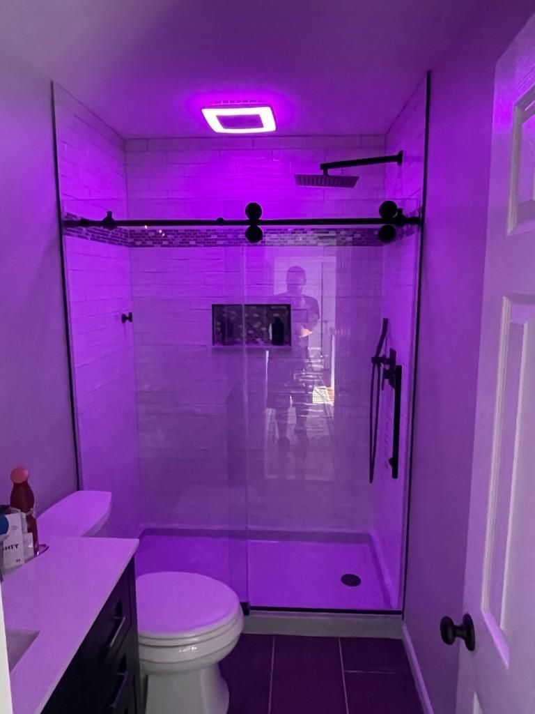 Bathroom with a glass-enclosed shower, toilet, and vanity under purple lighting.