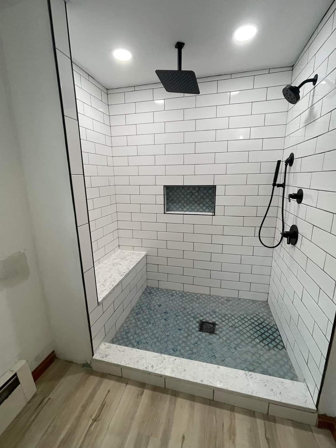 Modern shower with white brick-style tile, black fixtures, built-in bench, and blue-gray floor tiles.
