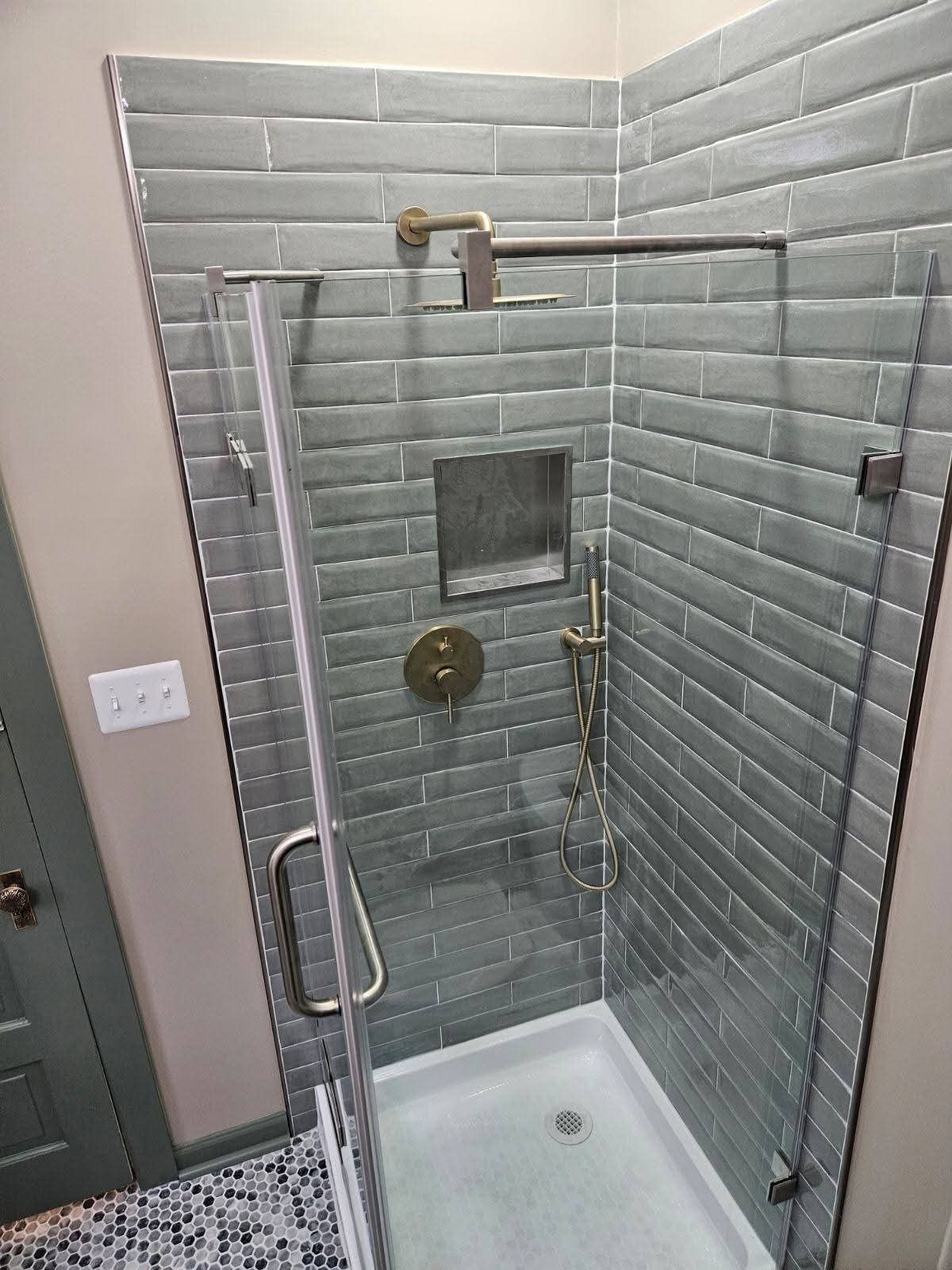 Small shower with gray tiled walls, glass door, and brass fixtures.