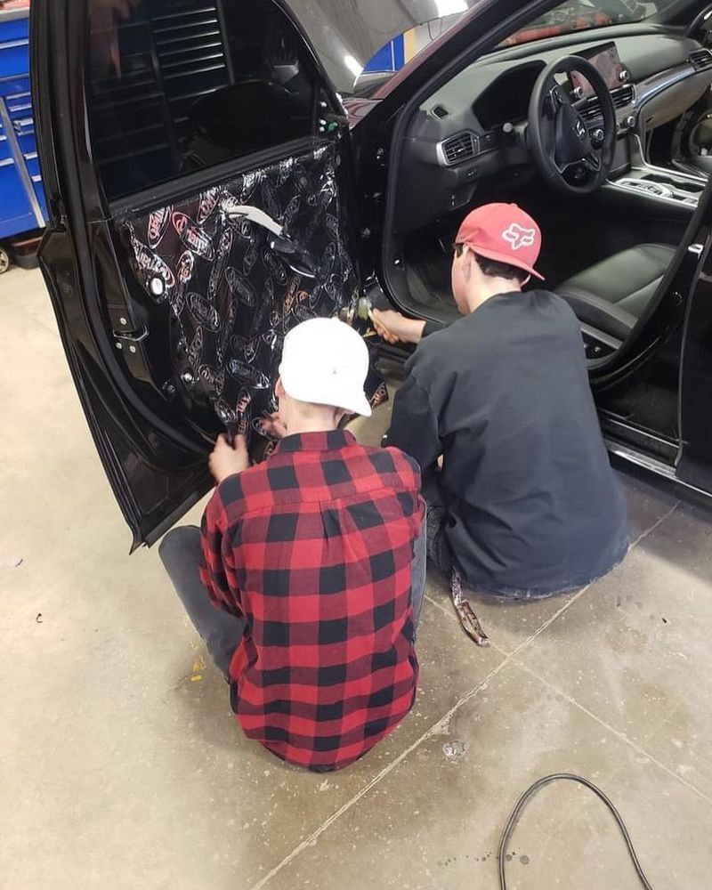 Two people working on a car door, one wearing a red and black plaid shirt, the other a red hat.