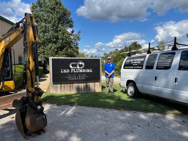 A man stands by a sign for C&D Plumbing, with a backhoe on the left and a white van on the right, under a blue sky.