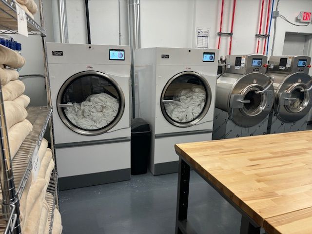 Commercial laundry room with two large white dryers in use; linens on shelves, and a wooden table.