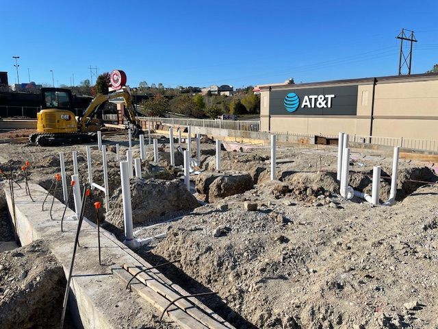 Construction site with plumbing pipes, an excavator, and an AT&T building under a blue sky.