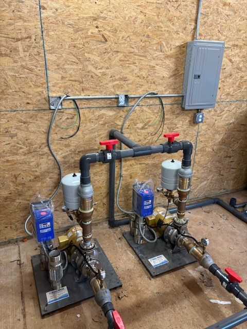Two water pumps with valves and a circuit breaker box mounted on a wooden wall.