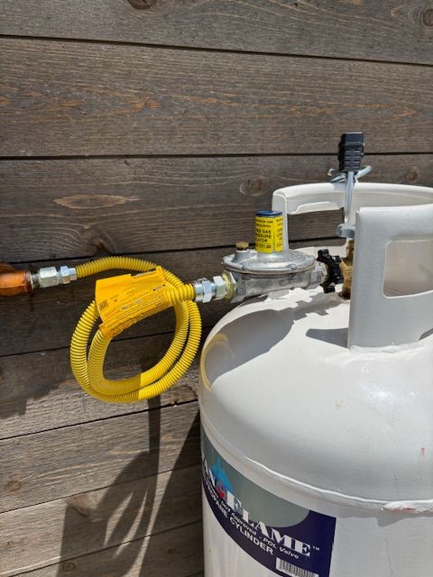 A propane tank with a yellow hose and regulator attached against a wood-paneled wall.