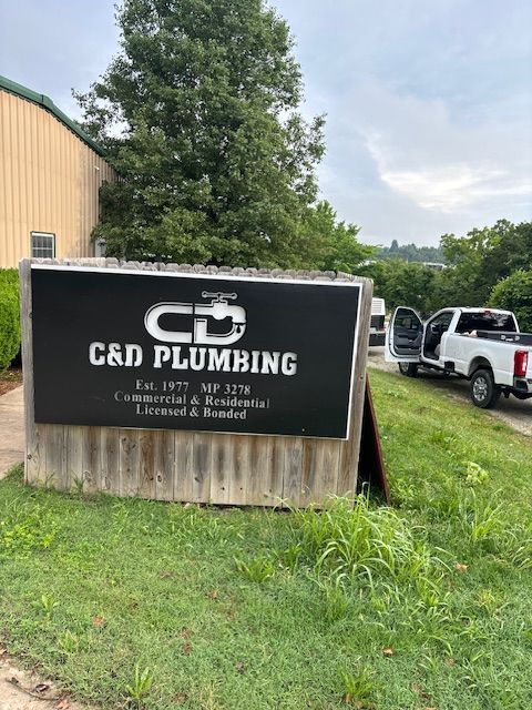 Sign for C&D Plumbing, on a cloudy day. White truck parked nearby. Building in background.