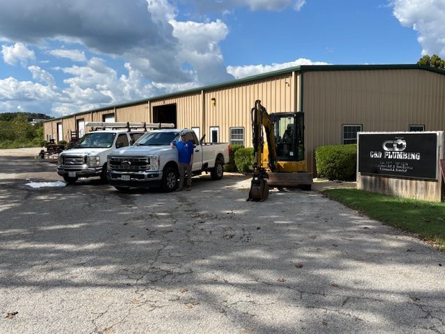 Vehicles and machinery in front of a tan building with a sign. A person stands near a white truck on a paved lot.