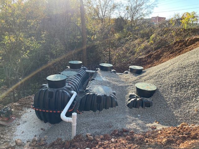 A septic system with several black tanks and access ports on a gravel bed, set against a hillside with trees.