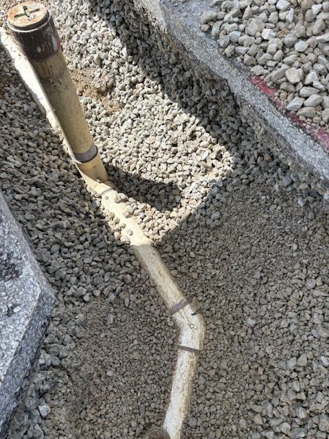 A pipe in a gravel-lined trench, near a concrete edge, angled in sunlight.