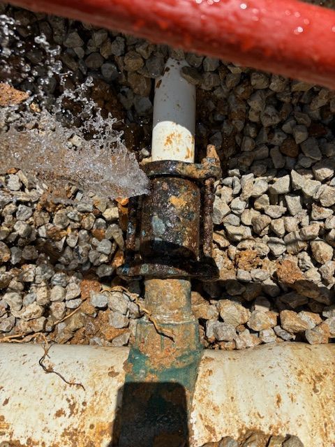 Water spraying from a corroded pipe joint surrounded by rocks and dirt.